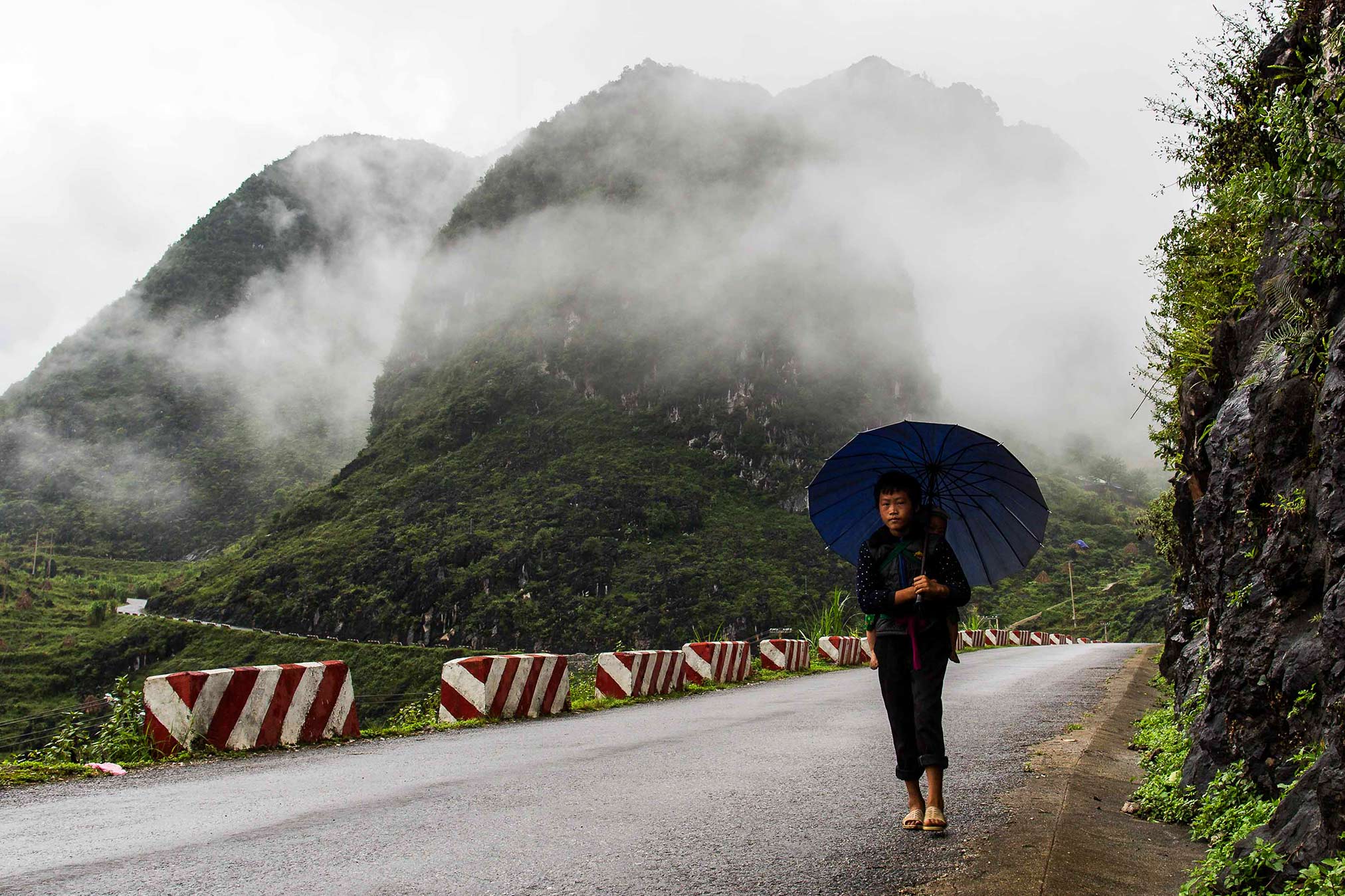Young Hmong woman walking on the road sheltering her child with a big blue umbrella